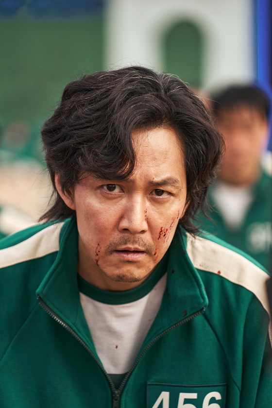 Actor Lee Jung-jae wearing a green tracksuit with blood splatters on his face, staring intensely forward in a scene from Squid Game