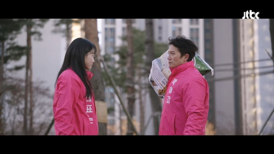 The main leads of the Korean drama The Apartment Job wearing matching pink jackets, facing off and confronting each other with tense expressions inside the apartment complex courtyard